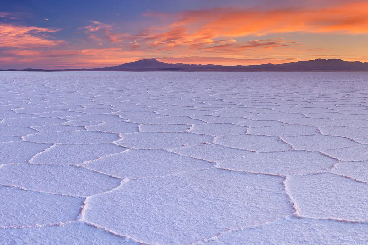 A large, textured salt flat with polygonal patterns stretches to a distant mountain range under a vibrant orange and blue sunset sky