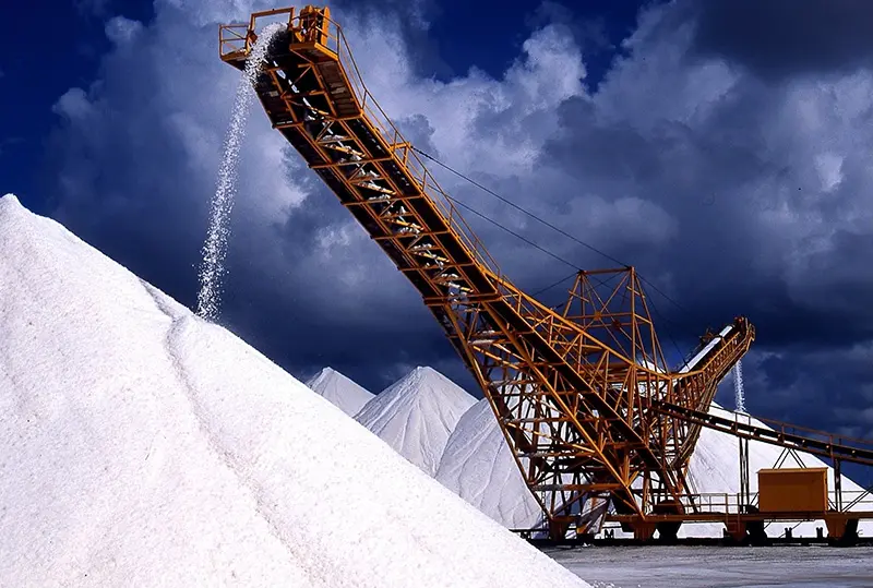 Several enormous piles of gleaming white salt surround an industrial yellow salt elevator