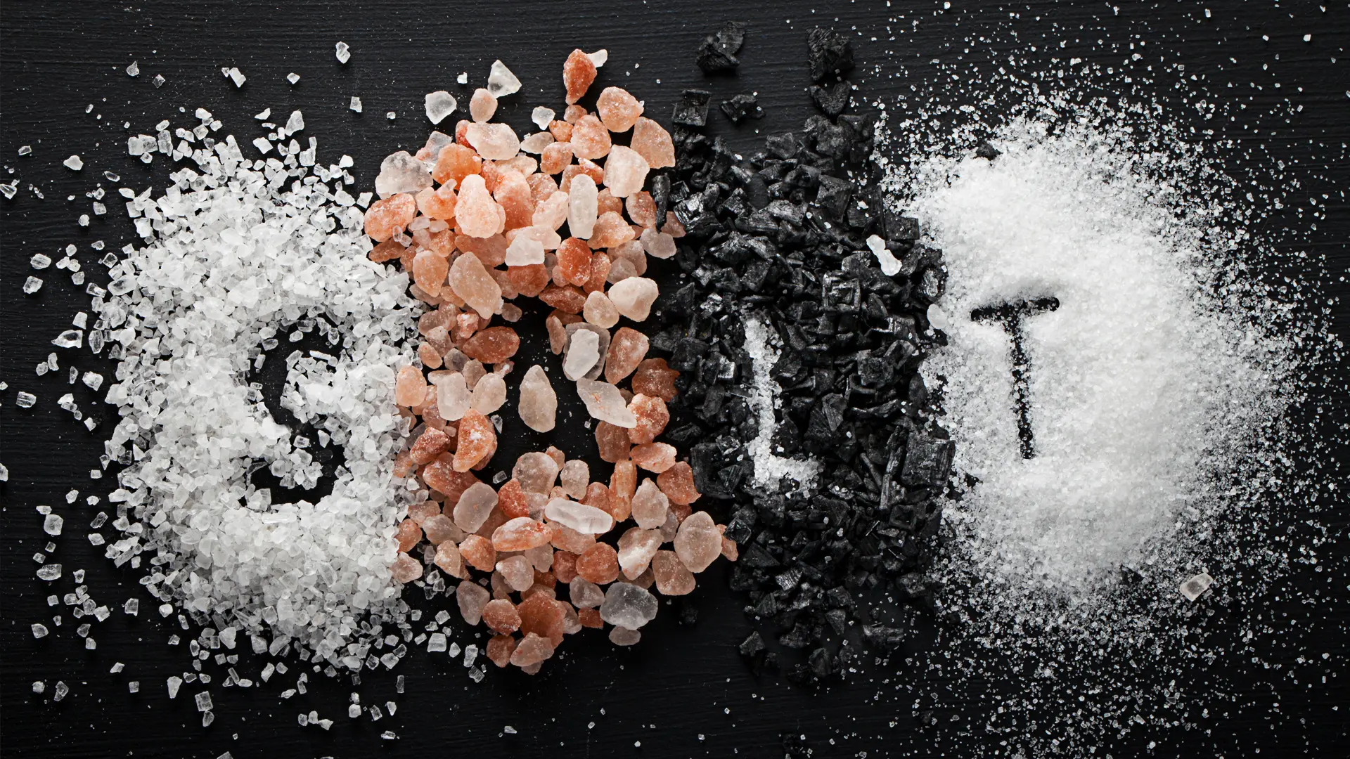 The word ‘SALT’ is spelled out using four different types of salt on a dark background: coarse white salt for ‘S’, pink Himalayan salt for ‘A’, black salt for ‘L’, and fine white salt for ‘T’